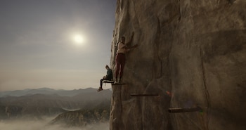 Una mujer de pie y otra sentada sobre plataformas de metal fijadas a un acantilado, con montañas y el sol de fondo