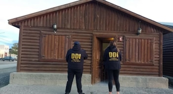 Dos agentes de policía con chaquetas de la DDI Santa Cruz frente a una cabaña de madera con la puerta abierta y el número uno