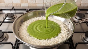 Jarra de vidrio que vierte salsa verde en el centro de una sartén con arroz blanco crudo, ubicada sobre los quemadores de una estufa de gas.