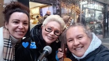 Tres mujeres sonrientes en primer plano tomando una selfie; una de cabello rubio platino sostiene un micrófono y otra un instrumento de cuerda, en una calle