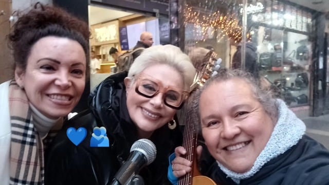 Carmen Barbieri posa sonriente junto a una artista callejera y otra mujer, con un micrófono y un instrumento musical, en una calle concurrida del centro de Buenos Aires.