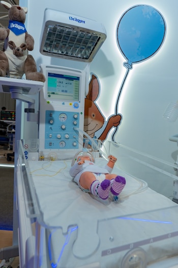 Equipo médico Dräger con pantalla, maniquí de bebé en incubadora con cables y mascarilla, peluche de canguro y decoraciones de pared