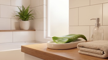 Ilustración digital de una hoja de aloe vera cortada mostrando su gel claro en una bandeja, junto a toallas y un dispensador de jabón en un baño blanco y madera.