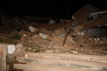 Escombros de una edificación destruida con techo desprendido, rodeada de tierra y algunos arbustos, junto a unas escaleras de cemento, de noche