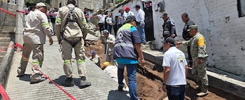 Un grupo de trabajadores y personal uniformado se congrega alrededor de una zanja abierta en una calle de concreto, con cinta roja de "peligro" visible