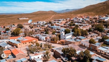 Vista aérea de casas bajas con techos de chapa, una iglesia blanca y calles de tierra rodeadas por cerros áridos bajo un cielo despejado