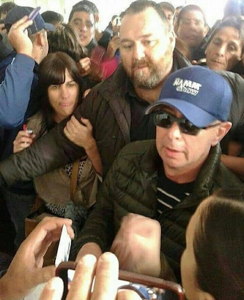 Un hombre con barba y chaqueta oscura, Diego Biscione, abre paso para otro hombre, El Indio Solari, que lleva gorra azul, gafas de sol y chaqueta oscura, entre una multitud