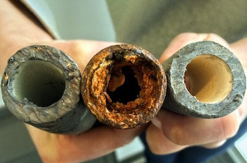 Unas manos sostienen tres secciones de tuberías de metal vistas de frente, de las cuales la del centro presenta óxido en su interior