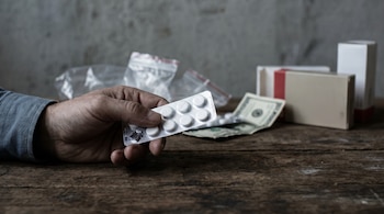 Mano adulta sosteniendo un blíster de pastillas genéricas parcialmente vacío. Sobre una mesa de madera se ven billetes, bolsas y cajas de medicamentos borrosas al fondo.