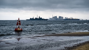 Una boya roja y blanca en el agua cerca de la playa, con un buque de guerra e instalaciones industriales al fondo bajo un cielo nublado.