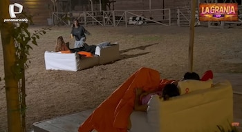 Shirley Arica de pie en un set exterior nocturno de "La Granja VIP", con otras personas relajándose en pufs y cubiertas con mantas, el suelo es de paja
