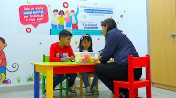Una mujer y dos niños sentados a una mesa de colores con un contenedor de juguetes y carteles informativos al fondo.