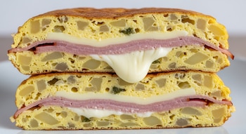 Una tortilla de papa gruesa cortada al medio, con capas de jamón cocido y queso cremoso derretido entre las mitades de la tortilla. Fondo blanco.