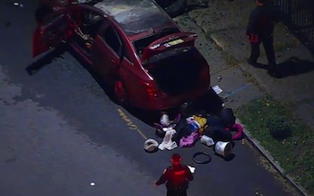 Vista aérea de un automóvil rojo con las puertas y el maletero abiertos, ropa y objetos en la calle, y un policía uniformado de espaldas