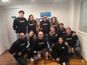 Un grupo de trece personas, ocho hombres y cinco mujeres, posa en una oficina con vestimenta negra de la marca Oceanic. Detrás se ve el logo de Oceanic en la pared y una puerta