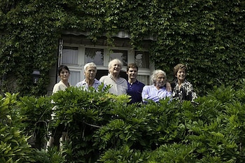 Cinco personas, tres hombres mayores, un hombre joven y dos mujeres, posan detrás de un seto verde con un edificio cubierto de hiedra