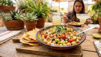 Primer plano de un tazón colorido de ceviche de garbanzos con tomate, cebolla, pepino y jalapeños, acompañado de nachos en una mesa de madera. Una persona come en el fondo