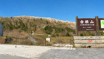 Letrero de madera sobre base de piedra con texto en japonés junto a un sendero y una colina con escasa vegetación bajo el cielo azul