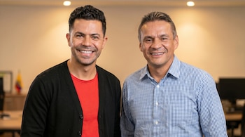 Dos hombres sonríen directamente a la cámara. Uno lleva una camiseta roja y cárdigan negro, el otro una camisa de cuadros azules y blancos.