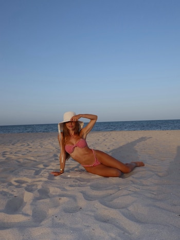 Mujer joven en bikini rojo a cuadros y sombrero blanco recostada en la arena de una playa. Al fondo, el mar y un cielo despejado