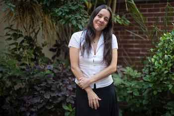 Retrato de Valeria Castro, mujer de cabello oscuro, con camisa blanca y falda negra, sonriendo a cámara, rodeada de plantas y una pared de ladrillo