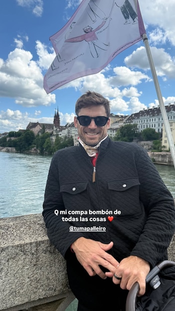 Hombre sonriente con gafas de sol, chaqueta negra y pañuelo, parado en un puente con un río y edificios históricos de una ciudad suiza de fondo