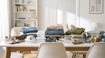 Mesa con pilas de ropa, pares de zapatos, cajas de medicamentos, tuppers plásticos, cables, revistas, utensilios y juguetes de madera.
