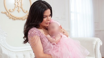 Ely Yutronic, con vestido rosa brillante, abraza a su hija Luna, cuyo rostro está difuminado, ambas vestidas de rosa, en un sofá blanco con fondo claro.