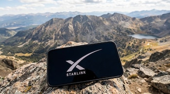 Un teléfono celular negro con el logo blanco de Starlink en la pantalla, apoyado sobre una roca. Al fondo, un vasto paisaje montañoso y un lago.