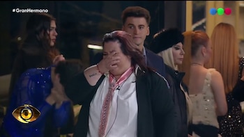Mujer de cabello rojizo con camisa blanca y corbata rosa se cubre el rostro con la mano y llora. Hay otras personas en el fondo y el logo de Gran Hermano