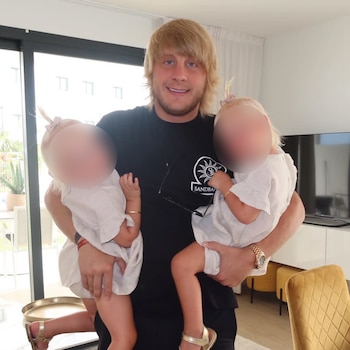 Hombre rubio con barba cargando a dos niños pequeños de cara borrosa; fondo con ventana, televisión, mueble blanco y silla amarilla