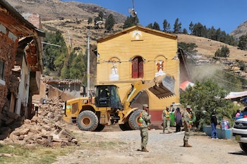 Máquina cargadora frontal amarilla retira escombros frente a un templo amarillo con fisuras. Tres soldados observan, con montañas y casas al fondo