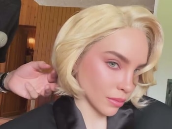 Mujer rubia de cabello ondulado y corto con blazer negro, su peinado es ajustado por una mano. Rostro de un hombre barbudo y pared de madera al fondo
