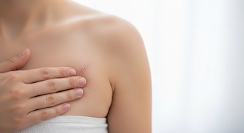 Primer plano de una mano femenina sobre el pecho de una persona con una cicatriz quirúrgica. La persona lleva una prenda blanca. Fondo blanco difuso.