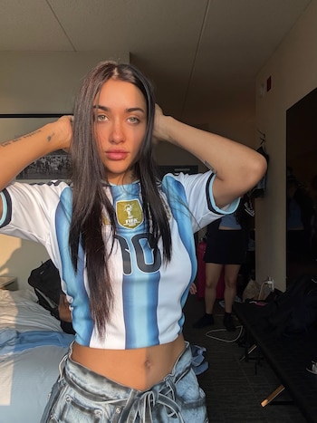 Mujer de cabello oscuro, ojos claros, camiseta argentina número 10 con parche FIFA World Cup 2022, pantalones de mezclilla, manos en la cabeza