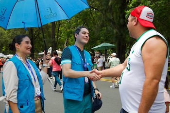 Tres personas en primer plano. Una mujer sostiene una sombrilla azul con el logo de Poder Ciudadano. Dos hombres se dan un apretón de manos. Hombre de gorra Texaco