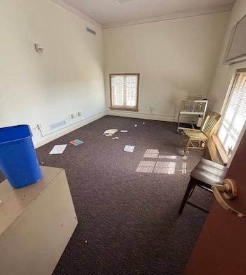 Una habitación con moqueta oscura, paredes claras, dos ventanas, dos sillas, un mueble bajo y papeles dispersos por el suelo