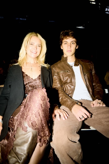 Una mujer rubia con abrigo negro y un joven con chaqueta de cuero marrón están sentados sonriendo ante la cámara