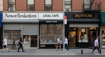 Fachada de varios locales comerciales en Nueva York, algunos con persianas metálicas cerradas u otras de rejilla, y personas caminando por la acera.