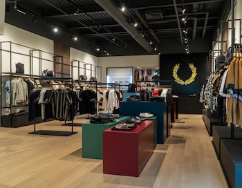 Interior de una tienda con estantes metálicos negros, ropa colgada, mesas expositoras de colores, zapatos, bolsos, y un logo dorado en una pared