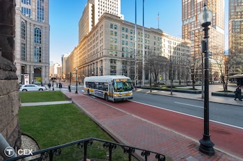Vista diurna de un autobús blanco y amarillo de la MBTA circulando por una calle roja dedicada, flanqueado por edificios, árboles y peatones en una ciudad