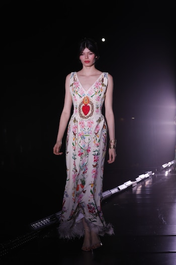 Modelo con vestido blanco de motivos florales y un corazón rojo bordado en el pecho camina por una pasarela oscura con luces