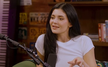 Kylie Jenner de frente, con pelo oscuro, camiseta blanca y maquillaje sutil, sentada frente a un micrófono negro en un estudio con placas de coche de fondo