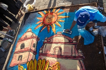 Un hombre con gorra azul crea una alfombra de aserrín colorida en el suelo, con diseños de un sol radiante y dos estructuras arquitectónicas coloniales, en una calle empedrada