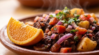 Un plato de barro contiene sangrecita cocida con trozos de papa, tomate y cebolla morada, junto a un trozo de naranja fresca.