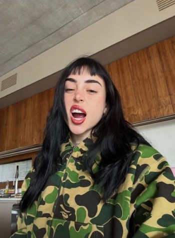 Mujer con cabello negro largo y flequillo, boca abierta con labios rojos, usa buzo camuflado verde y negro. Fondo interior con madera