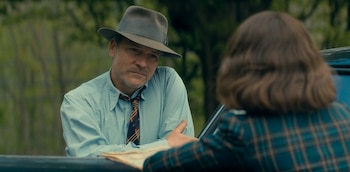 Peter Sarsgaard, con sombrero gris y camisa azul, se inclina y mira a una persona de espaldas con cabello castaño y chaqueta a cuadros. Fondo verde