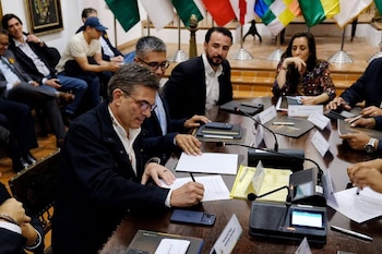 Hombre con gafas y chaqueta oscura firma documento en mesa de madera oscura. Se observan personas, papeles, micrófonos y banderas de colores
