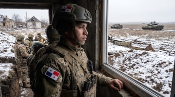 Primer plano de joven militar con uniforme camuflado, casco y bandera de Panamá en el parche. Mira por una ventana hacia un campo nevado con tanques y ruinas.