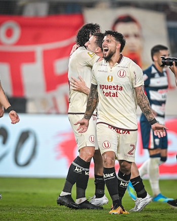 Tres futbolistas de Universitario con camiseta crema celebran en el campo. El jugador en primer plano grita con los brazos abiertos, mientras otros dos se abrazan detrás